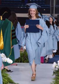 A smiling young woman holding her graduation certificate walks in a line of fellow grqaduates